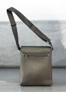 A black pebbled leather crossbody bag with a wide adjustable shoulder strap, standing upright on a gray surface against a light gray background. By Fresh N Fitted.