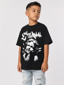 A young boy wearing a black Stencil band graphic t-shirt with a white stylized logo and a black-and-white portrait print, paired with light-wash ripped jeans, against a light gray studio background.
