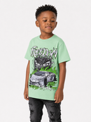 A young boy wearing a light green graphic t-shirt with a black and white car and cat design, paired with distressed black jeans, standing against a light gray studio background.