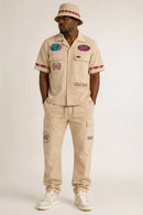 A man modeling a beige short-sleeve button-up shirt and matching cargo pants set with embroidered patches and red stripe accents, paired with a beige hat and white sneakers, against a light gray studio background. By Fresh N Fitted
