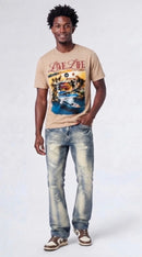 A man modeling a beige graphic t-shirt with a tropical resort scene, paired with distressed blue jeans and brown sneakers, against a light gray studio background. 