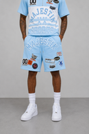 A model wearing a light blue Majestic Sports two-piece tracksuit, featuring a short-sleeve t-shirt and matching shorts, both decorated with white graphic prints and various colorful patches. The model is also wearing white socks and white sneakers, standing against a neutral gray studio background.