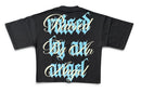 A black short-sleeve t-shirt shown from the back, featuring a large graphic print with the text 'Raised By An Angel' in stylized white and light blue gothic lettering. By Fresh N Fitted.