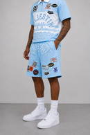 A model wearing a light blue and white two-piece tracksuit with various graphic patches and text, paired with white socks and white sneakers, set against a neutral gray studio background.