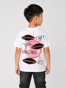 A child model shown from the back wearing a white short-sleeve t-shirt with a graphic print of four black UFOs, a pink circular scribble, and the text 'THEY ARE AMONG US'.