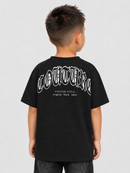 A child viewed from the back wearing a black short-sleeve T-shirt with a large white gothic-style graphic and text across the upper back, paired with dark jeans, against a light gray studio background.