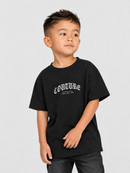 A young boy wearing a black short-sleeve t-shirt with a white gothic-style 'COUTURE' graphic on the chest, paired with dark gray jeans, photographed against a light gray studio background.