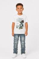 A young boy modeling a white graphic t-shirt with a green cartoon character and text, paired with distressed blue jeans and white sneakers, against a plain light gray background.