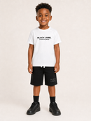 A young boy stands against a light beige studio background wearing a white t-shirt with 'BLACK LABEL' printed in black on the chest, black shorts with a matching print on the leg, black socks, and black sneakers.