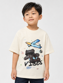 A young boy wearing a cream-colored short-sleeve t-shirt with a graphic of a blue airplane, a black teddy bear, and the word 'VISES' in black script, set against a plain light gray background.