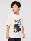 A young boy wearing a cream-colored short-sleeve t-shirt with a graphic of a black teddy bear, a blue airplane, and the word 'VISORS' in black text, paired with dark blue jeans, photographed against a light gray studio background.