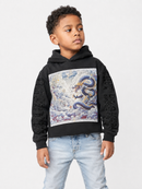A young boy wearing a black hoodie with a large graphic print of a blue and gold dragon on the front, paired with light wash jeans, photographed against a light gray studio background.