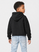 A child model shown from the back wearing a black cropped hoodie with textured lace-like sleeve panels and light blue jeans, photographed against a plain light gray studio background.