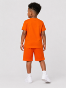 A young boy viewed from behind wearing a bright orange short-sleeve t-shirt and matching orange athletic shorts, paired with white socks and white sneakers, against a light gray studio background.