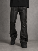 Black leather flare jeans with raised gothic-style lettering and flame-like embroidery details on the thighs, shown on a model from the waist down against a textured gray studio background.