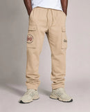 A person wearing beige cargo jogger pants with side flap pockets, a drawstring waist, and embroidered logos on the left thigh and right hip, paired with light-colored sneakers against a plain studio background. By Fresh N Fitted