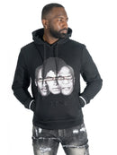 A man wearing a black hoodie with a large black-and-white graphic print of three faces and the text 'GREATEST OF ALL TIME' across the chest, paired with distressed gray jeans, against a plain white background.