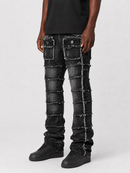 A person wearing heavily distressed black denim jeans with stacked patchwork panels and frayed seams, paired with black shoes against a light gray studio background.