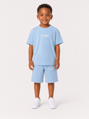 A young boy modeling a light blue two-piece set consisting of a short-sleeve t-shirt and matching shorts, both featuring the word 'Saint' printed in white Gothic lettering. He is standing against a clean, off-white studio background.