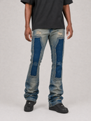 A person wearing distressed light-wash blue jeans with large dark blue patch panels on the thighs, shown from the waist down against a light gray studio background.