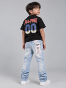 A young boy is shown from the back wearing a black t-shirt with a large graphic on the back, light-wash blue jeans with a patch on the back pocket, and white sneakers, set against a plain gray studio background.