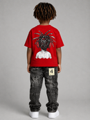 A child viewed from behind wearing a bright red short-sleeve T-shirt with a large graphic print of a person with dreadlocks on the back, paired with dark washed jeans and white sneakers, against a plain gray studio background.