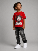A young boy with dreadlocks stands against a gray studio background wearing a bright red graphic t-shirt, distressed black jeans, and white sneakers. The t-shirt features a graphic of a doll with red hair and a red mouthpiece.