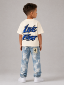A young boy is shown from the back wearing a cream-colored t-shirt with a blue 'LIVE FAST' graphic print, light blue distressed jeans, and white sneakers, set against a plain gray studio background.