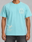 A man wearing a light blue graphic t-shirt with white printed designs on the chest, shoulders, and lower front, paired with dark gray jeans against a neutral gray background. By Fresh N Fitted