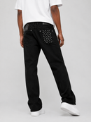 Rear view of a person wearing black jeans with decorative silver hardware on the back pockets, paired with a white t-shirt and white sneakers, against a light gray studio background.