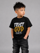 A young boy wearing a black graphic t-shirt with 'TRUST GOD DSATR' printed on the front, paired with dark gray jeans, standing against a solid gray studio background.