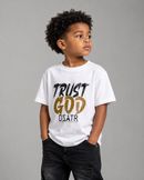 A young boy with curly black hair wearing a white crewneck t-shirt with a black and gold graphic that reads 'TRUST GOD DSATR', paired with black jeans, against a solid gray studio background.