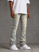 A person wearing light blue and beige distressed denim jeans with a faded abstract pattern, paired with a white t-shirt and white sneakers, standing against a textured gray studio background.