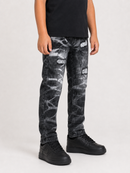 A child wearing distressed black and gray acid-wash jeans with white graphic patches, a black t-shirt, and black sneakers, shown from the waist down on a light gray studio background.
