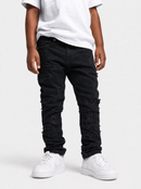 A child wearing black heavily distressed ripped jeans, a white t-shirt, and white sneakers, standing against a light gray studio background.