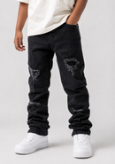 A child wearing black distressed denim jeans with ripped details on the thighs and knees, styled with a white t-shirt and white sneakers against a light gray studio background.