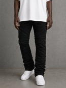 A person wearing black distressed denim jeans with diagonal frayed cut details, paired with a white t-shirt and white sneakers, standing against a textured gray wall.