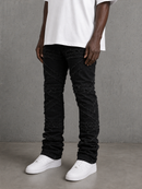 A person wearing black distressed jeans with textured, frayed paneling down the legs, styled with a white t-shirt and white sneakers against a gray concrete wall.