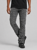 A person wearing faded black denim jeans with diagonal distressed patchwork panels and frayed detailing, paired with a black t-shirt and black sneakers, against a light gray studio background.
