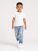 A young boy walking forward in a studio setting, wearing a plain white short-sleeve t-shirt, light blue distressed denim jeans, and white sneakers against a light gray background.