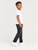 A young boy standing in profile wearing a white short-sleeve t-shirt, dark gray speckled jeans, and white sneakers against a light gray studio background.