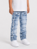 A child wearing light blue heavily distressed patchwork denim jeans with frayed edges, paired with a white t-shirt and white sneakers, shown from the waist down against a light gray studio background.