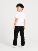 A young boy standing in profile wearing a plain white t-shirt, black distressed jeans with layered ruffle details, and white sneakers against a light gray studio background.