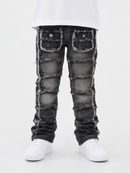 A pair of distressed black denim jeans with patchwork paneling and frayed edges, shown on a model from the waist down wearing a white t-shirt and white sneakers against a white studio background.