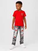 A young boy modeling a bright red short-sleeve t-shirt with a small red graphic on the left chest, paired with distressed gray jeans featuring red ripped details, and white sneakers, against a light gray studio background.