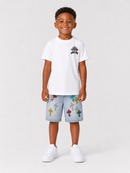 A young boy stands against a light gray studio background wearing a white short-sleeve t-shirt with a small multicolor cross graphic on the left chest, light-wash denim shorts covered with colorful embroidered crosses, and white sneakers.