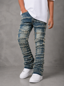 A person wearing distressed blue denim jeans with heavily ripped and frayed detailing, styled with a white t-shirt and white sneakers against a gray studio background.