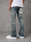 Rusticated blue denim flare jeans with heavily distressed, gathered legs, shown on a model from the back against a gray concrete wall.