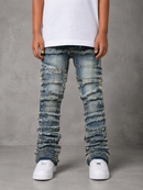 A person wearing heavily distressed and frayed blue denim jeans with a ripped, layered look, styled with a white t-shirt and white sneakers against a gray textured studio background.