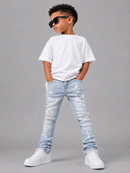 A child model wearing a white t-shirt, light-wash distressed denim jeans, white sneakers, and black sunglasses, standing against a solid gray studio background.